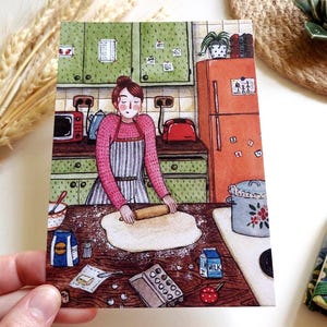 Könnte beinhalten: Aquarellillustration einer Frau in einem rosa Pullover und einer gestreiften Schürze, die in einer Küche Teig ausrollt. Die Küche hat grüne Schränke, einen orangefarbenen Kühlschrank und Kochutensilien. Mehl, Milch und andere Zutaten auf dem Tisch.
