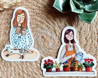 Może przedstawiać: Dwie naklejki z ilustracjami kobiet. Jedna czyta książkę ze słowem "Tales" widocznym i kotem. Druga podlewa rośliny na półce. Naklejki są w stylu akwareli.