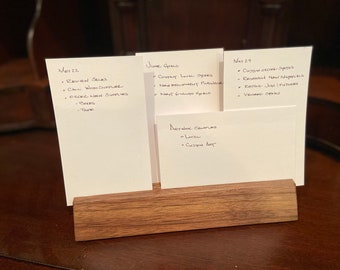 Könnte beinhalten: Ein Holzständer mit fünf weißen Karteikarten, jede mit handschriftlichen Notizen für die Monate Mai, Juni, September und November. Die Notizen listen Aufgaben und Erinnerungen für jeden Monat auf.