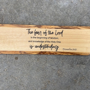 May include: Wooden bookmark with a light brown wood grain and a dark brown wood border. The bookmark has a quote from Proverbs 9:10 that reads "The fear of the Lord is the beginning of wisdom, and knowledge of the Holy One is understanding."