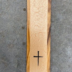 May include: A wooden cutting board with a cross design burned into the surface. The board is made of two different types of wood, one light and one dark.