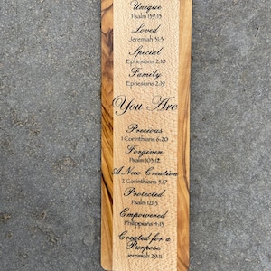 May include: Wooden bookmark with a light brown wood background and dark brown wood trim. The bookmark has a laser-engraved design with the text "You Are" in a large font and a list of words and bible verses below. The words include "Unique", "Loved", "Special", "Family", "Precious", "Forgiven", "A New Creation", "Protected", "Empowered", and "Created for a Purpose".