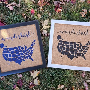 以下が含まれることがあります： 「wanderlust」という言葉が上に筆記体で書かれた、額縁付きのコルクボードが2つあります。ボードには、各州が輪郭で描かれたアメリカ合衆国の地図が描かれています。1つのボードは黒い額縁と白い州の輪郭で描かれた黒い地図です。もう1つのボードは白い額縁と白い州の輪郭で描かれた青い地図です。