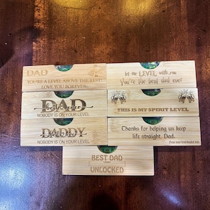 May include: A collection of bamboo spirit levels with engraved text. The levels have green vials and various messages for fathers, including "DAD," "BEST DAD," and phrases like "You're the best dad ever."