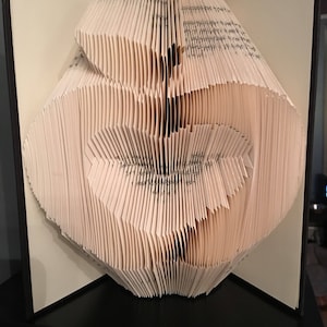 May include: A book folded into the shape of a heart. The pages are white and the book is closed. The book is sitting on a black surface.