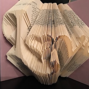 Puede incluir: Una escultura de arte de libro, doblada en forma de la palabra "LOVE", con las páginas del libro visibles. La escultura se muestra sobre una superficie rosa.
