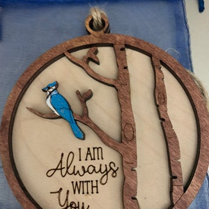 May include: A wooden ornament with a bluebird perched on a branch. The ornament is round and has a brown wood frame. The text "I am Always With You" is engraved on the ornament.