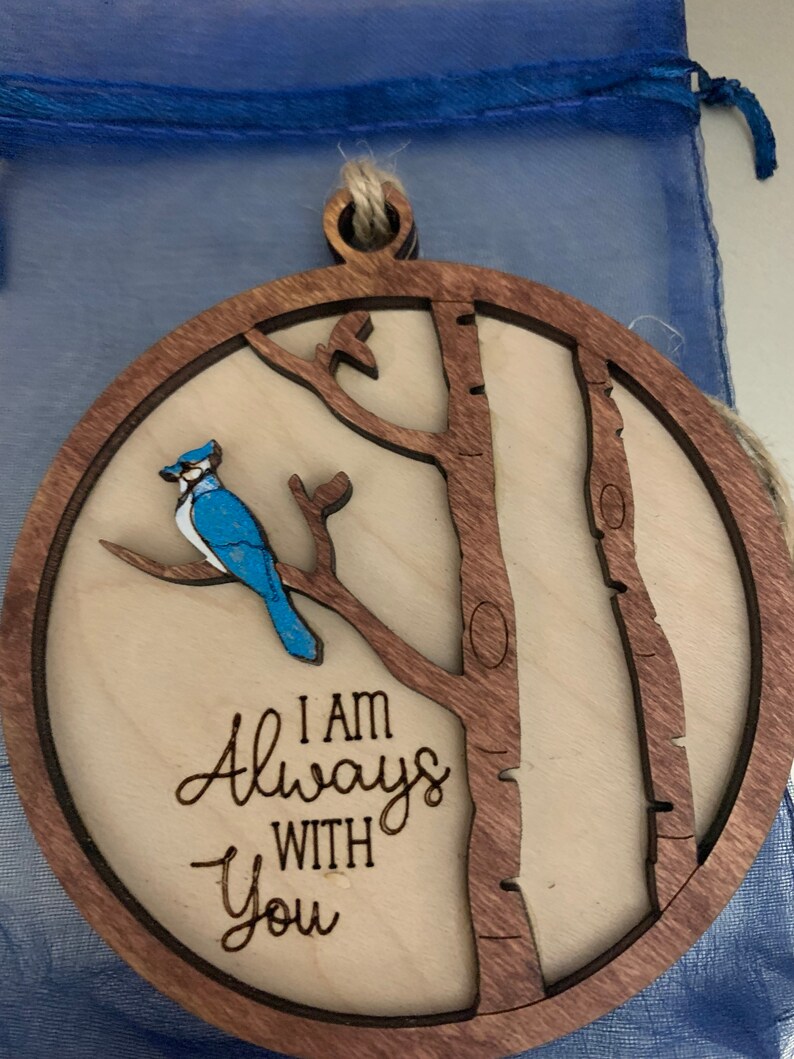 May include: A wooden ornament with a bluebird perched on a branch. The ornament is round and has a brown wood frame. The text "I am Always With You" is engraved on the ornament.