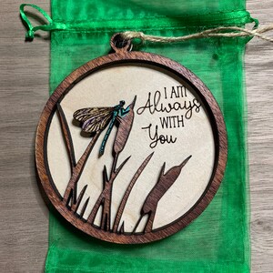 May include: A round wooden ornament with a cutout design of cattails and a dragonfly. The ornament is brown with a natural wood finish. The text "I am Always With You" is engraved on the ornament.
