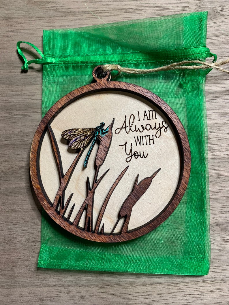 May include: A round wooden ornament with a cutout design of cattails and a dragonfly. The ornament is brown with a natural wood finish. The text "I am Always With You" is engraved on the ornament.