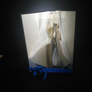 May include: A book folded into the shape of a bird, with a blue ribbon tied around it. The book is open and the pages are folded to create the shape of the bird.