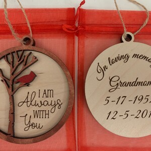 May include: Two wooden Christmas ornaments. The first ornament features a cardinal perched on a tree branch with the text "I am Always With You". The second ornament is a memorial ornament with the text "In loving memory of Grandmother 5-17-1952 - 12-5-2019".