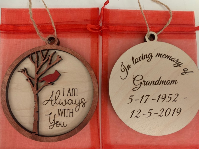 May include: Two wooden Christmas ornaments. The first ornament features a cardinal perched on a tree branch with the text "I am Always With You". The second ornament is a memorial ornament with the text "In loving memory of Grandmother 5-17-1952 - 12-5-2019".
