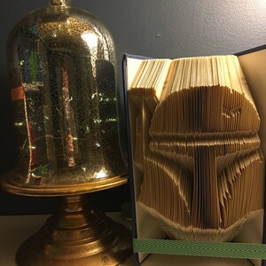 May include: A book art sculpture of a Star Wars character, Darth Vader, made by folding the pages of a book. The sculpture is displayed next to a gold-colored decorative stand with a glass dome.