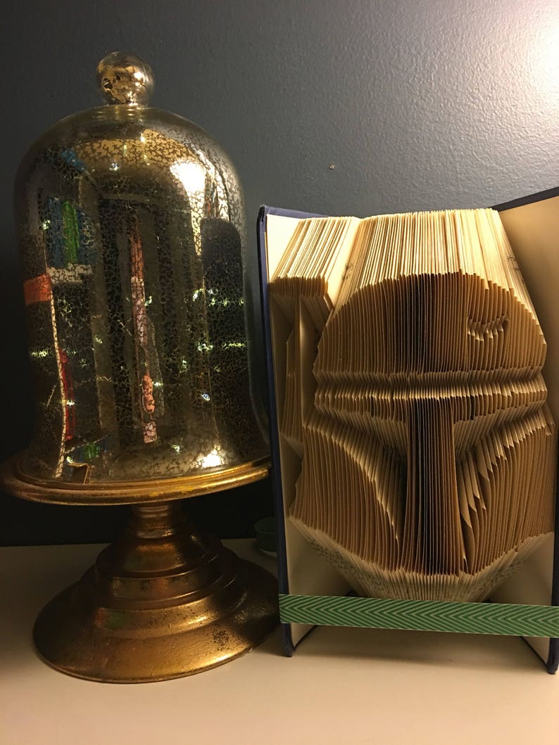 May include: A book art sculpture of a Star Wars character, Darth Vader, made by folding the pages of a book. The sculpture is displayed next to a gold-colored decorative stand with a glass dome.