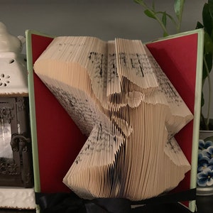 May include: A book art sculpture, folded into a three-dimensional shape, with a red and green cover and a black ribbon. The sculpture is on a black surface.