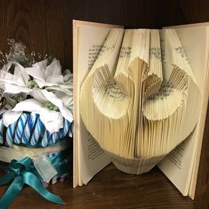 May include: A book with pages folded into a three-dimensional heart shape. The book is open and the heart shape is visible. The book is on a wooden shelf.