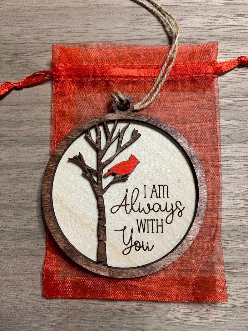 May include: A wooden ornament with a red cardinal perched on a tree branch. The ornament is round and has a brown wood frame. The text "I am Always With You" is engraved on the ornament.