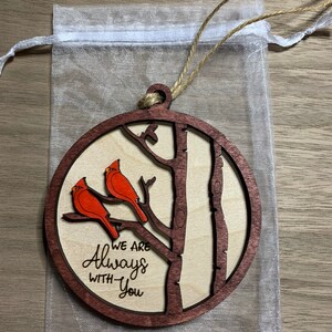 May include: A wooden ornament with a brown background and a tree with two red cardinals perched on branches. The text "We are Always With You" is engraved on the ornament.