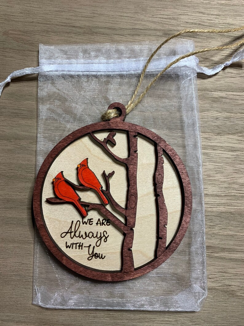 May include: A wooden ornament with a brown background and a tree with two red cardinals perched on branches. The text "We are Always With You" is engraved on the ornament.