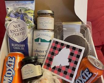 May include: A white gift box filled with Maine-themed goodies. The box contains a can of Moxie soda, a jar of Wild Maine Blueberry Jam, a can of Bar Harbor Lobster Chowder, a jar of Maine Seafood Rub, a bag of sweets, a blueberry cobbler mix, a coffee bag, and a red and black plaid coaster with a white outline of the state of Maine.
