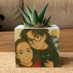 May include: A square concrete planter with a hand-painted illustration of two people and a black cat. The planter holds a green succulent plant. The planter sits on a rectangular cork board.