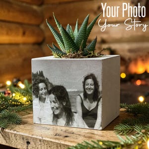 Op de afbeelding: Een vierkante betonnen plantenbak met een zwart-witfoto van drie personen op de voorzijde gedrukt. Een groene vetplant groeit uit de bovenkant van de plantenbak. De tekst Your Photo Your Story is hierboven zichtbaar.