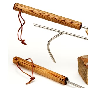 May include: Close-up of two metal fire pit tools with wooden handles and leather loops. The handles have a light brown wood grain pattern. The metal rods are silver. A piece of firewood is in the background.