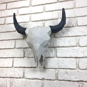 May include: A white paper mache bull skull with black paper horns mounted on a white brick wall.
