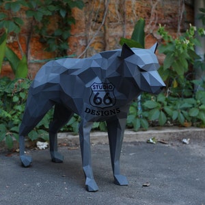 Könnte beinhalten: Eine graue Origami-Wolfskulptur mit einem schwarzen "Studio 66 Designs"-Logo an der Seite. Der Wolf steht auf einer grauen Asphaltfläche.