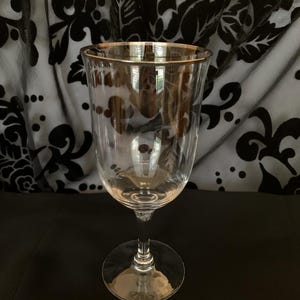 Puede incluir: Una copa de vino de vidrio transparente con un borde dorado. La copa tiene un tallo alto y delgado y una copa ancha.
