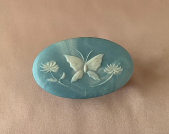 May include: An oval-shaped blue brooch with a marbled finish featuring a raised white relief design. The central motif depicts a butterfly flanked by two flowers, one on each side, with a smooth, polished surface and a distinct three-dimensional texture.