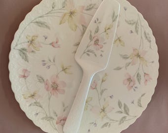 May include: A round white ceramic plate with a scalloped edge and a delicate floral pattern in soft pink, yellow, and green hues. A matching white ceramic cake server with a coordinating floral design rests on top of the plate.