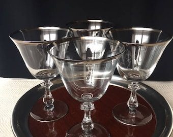 May include: Set of four clear glass stemmed drinking glasses with a silver rim. The glasses are on a brown tray with a silver rim.
