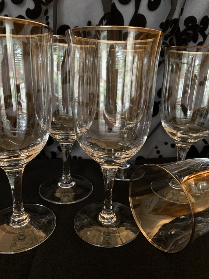 Puede incluir: Juego de seis copas de vino de vidrio transparente con borde dorado. Las copas est&aacute;n dispuestas en c&iacute;rculo sobre una superficie negra.