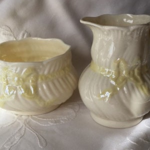 May include: A white ceramic creamer and sugar bowl set with yellow floral designs. The creamer has a handle and a spout. The sugar bowl is round and has a lid.