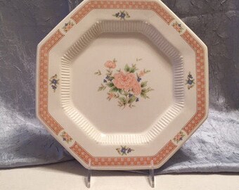 Puede incluir: Un plato octogonal blanco con un diseño floral. El plato presenta un borde a cuadros naranja y blanco. El centro del plato tiene un ramo de flores rosas y azules. El plato se exhibe sobre un soporte transparente.