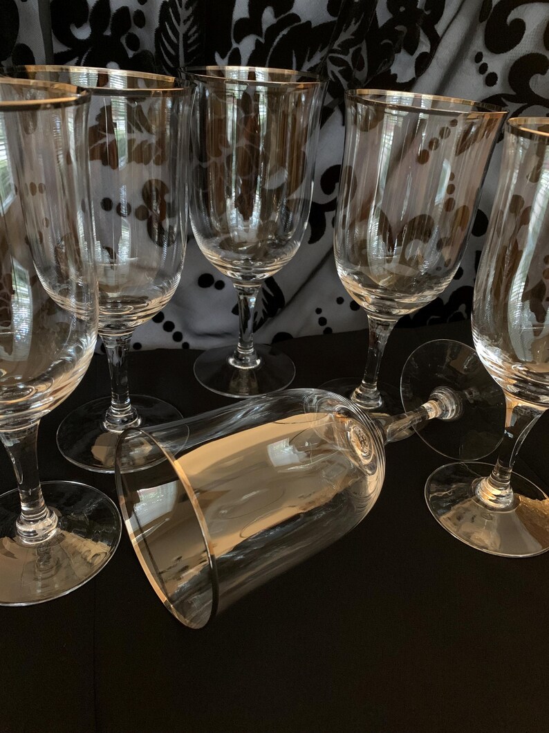 Puede incluir: Conjunto de seis copas de vino de vidrio transparente con borde dorado. Las copas est&aacute;n dispuestas en semic&iacute;rculo sobre una superficie negra.