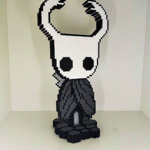 Pode incluir: Escultura em pixel art de um personagem do videogame Hollow Knight. A figura é principalmente branca e preta, com uma cabeça estilizada e grandes chifres. A figura está em uma base preta.