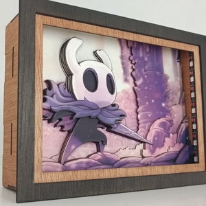 Peut inclure: Une boîte à ombre 3D représentant un personnage violet et blanc du jeu vidéo Hollow Knight. Le personnage tient une épée et se tient devant un fond violet et blanc.