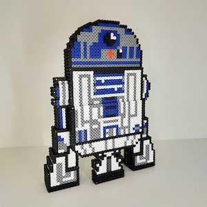 May include: A pixelated R2-D2 figure, crafted from small, colorful beads. The droid features a blue, white, gray, and black color scheme, with a rounded head and rectangular body. The figure stands upright, showcasing its iconic design.