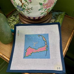 May include: Needlepoint canvas featuring a map of Cape Cod, Massachusetts, in pink and green against a blue background. The canvas is framed with a dark blue border. The canvas is on a woven surface, with a decorative ceramic vase in the background.