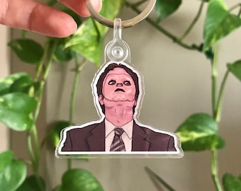 May include: A clear acrylic keychain featuring a cartoon illustration of a man with a surprised expression, wearing a dark suit and a patterned tie. The man is looking upwards.