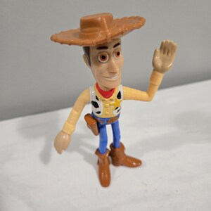 Pu&ograve; includere: Una figurina di plastica di Woody del franchise Toy Story. Woody indossa il suo cappello da cowboy marrone, la camicia gialla, il bandana rosso e i jeans blu. Sta alzando il braccio destro in segno di saluto.
