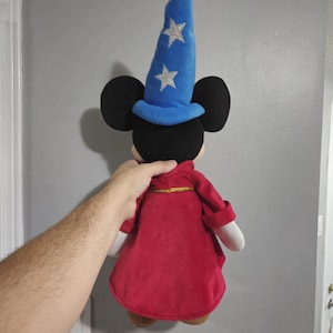 May include: A plush Mickey Mouse doll wearing a blue wizard hat with silver stars and a red robe with gold trim.