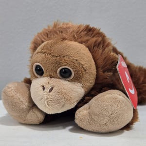 May include: A plush orangutan toy with brown fur and large, dark eyes. The stuffed animal has a light brown face and hands, and a red tag is attached. The toy is lying down.