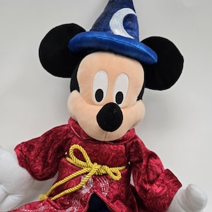 Pu&ograve; includere: Un Mickey Mouse di peluche che indossa una tunica rossa con bordi dorati e un cappello da mago blu con un design a mezzaluna.