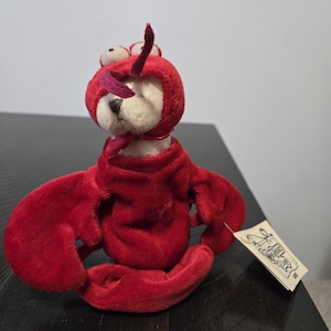 Könnte beinhalten: Ein Plüschtierbär in einem leuchtend roten Hummerkostüm. Der Bär hat ein weißes Gesicht und trägt eine rote Kapuze mit Wackelaugen und Antennen. Das Hummerkostüm hat große rote Scheren. Ein kleines Etikett ist angebracht.