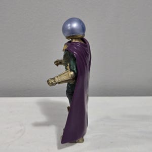 May include: A side view of a Mysterio action figure. The figure has a silver, spherical helmet, a purple cape, and gold-colored armor on the arms. The figure is standing on a white surface against a gray background.