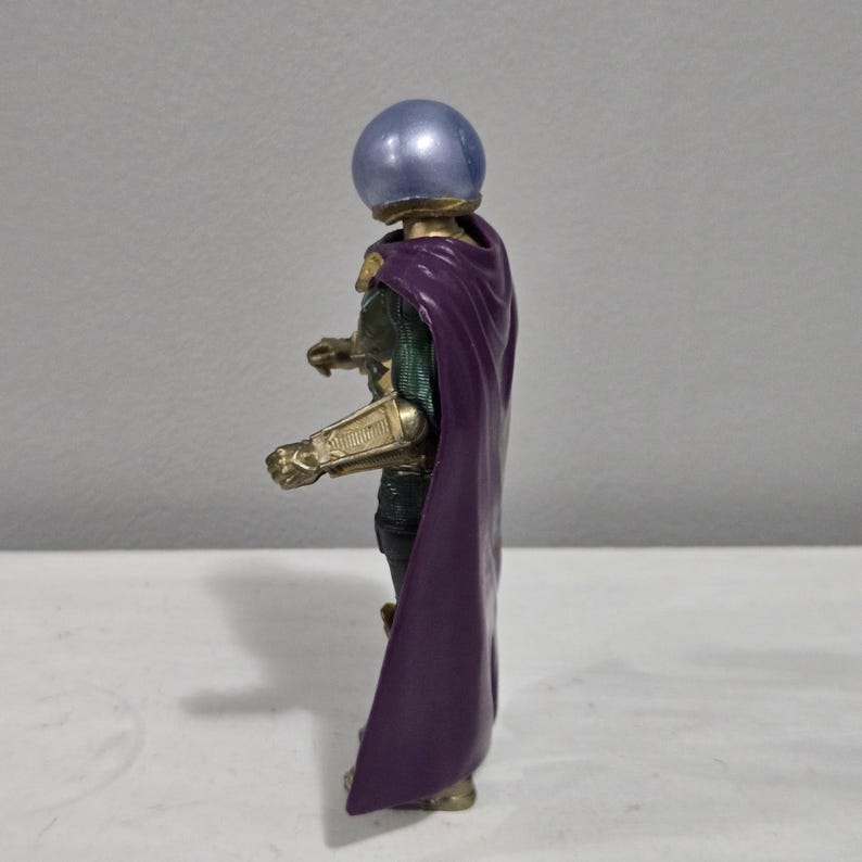 May include: A side view of a Mysterio action figure. The figure has a silver, spherical helmet, a purple cape, and gold-colored armor on the arms. The figure is standing on a white surface against a gray background.